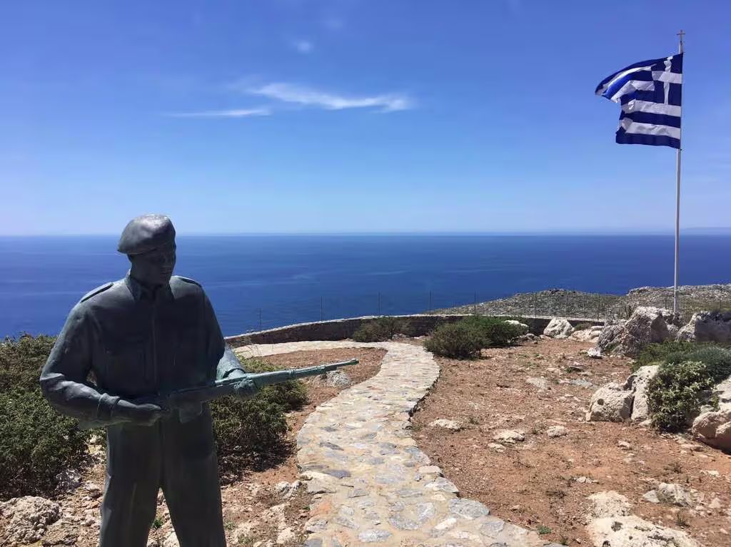crete_war2_tour A wartime soldier statue beside a Greek flag overlooking the deep blue Aegean Sea on a rugged coastal landscape in Crete.
