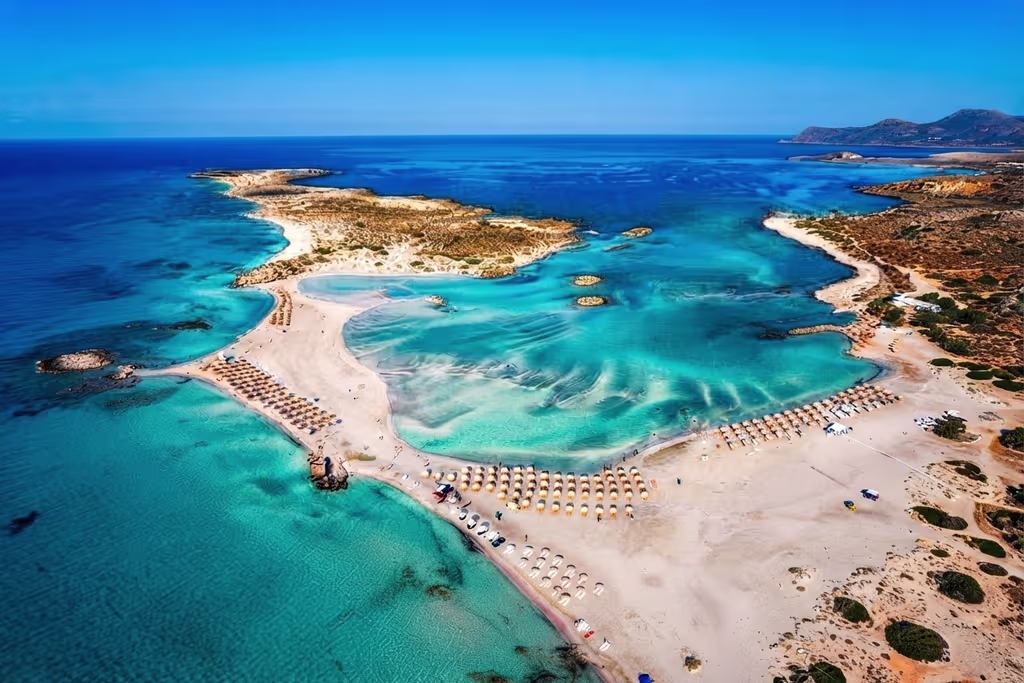 elafonissi_view Aerial view of Elafonissi Beach in Crete, showing turquoise lagoons, pale pink sandbars, rows of sun loungers and the surrounding rugged coastline.