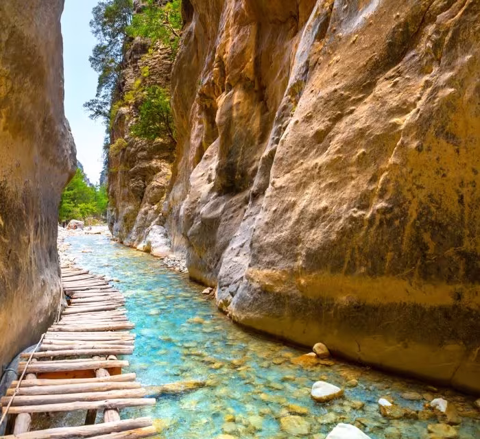 Wooden footbridge over a clear stream between the towering rock walls of Samaria Gorge in Crete, surrounded by lush vegetation.