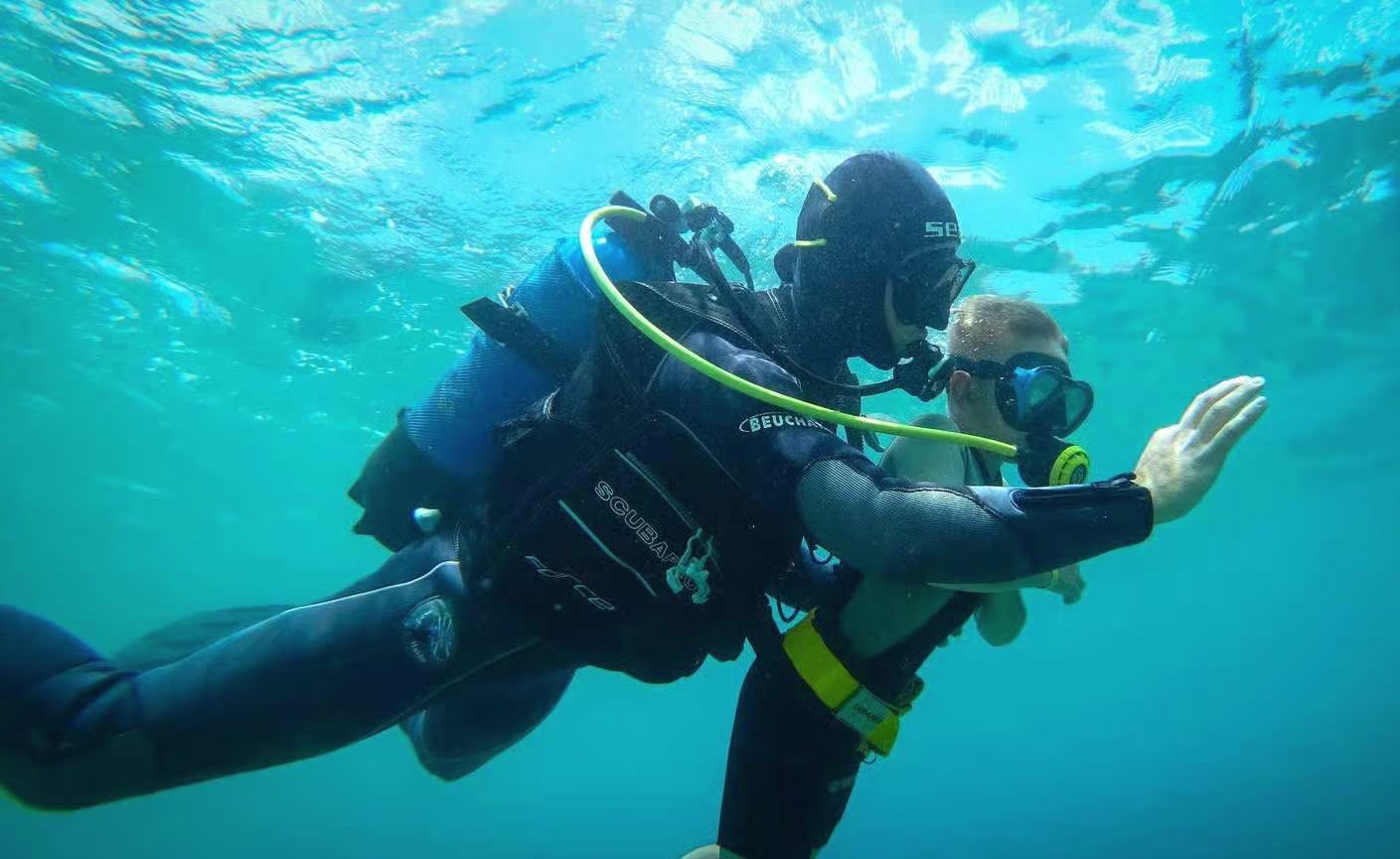 scuba heraklion (1) Two scuba divers exploring the clear blue waters off Heraklion, Crete, with one diver signaling underwater near the surface.
