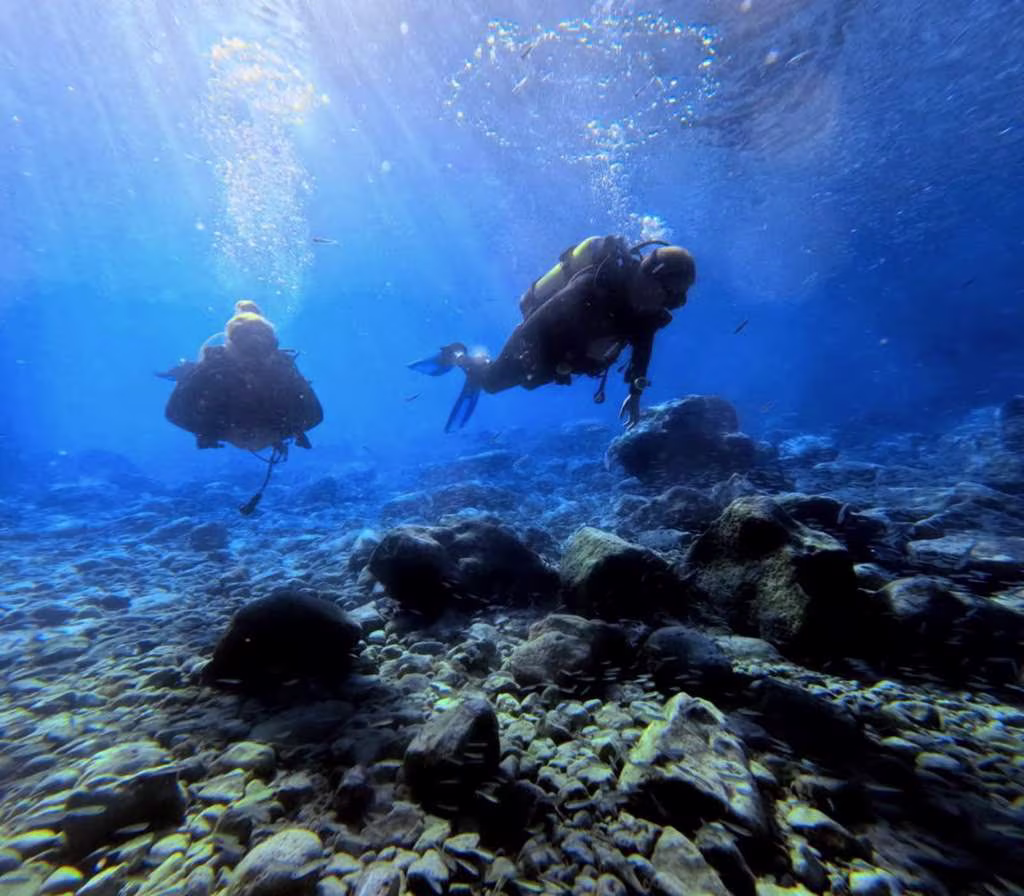 shore_dive_crete (1) Two scuba divers exploring the rocky seabed in the clear blue waters off Crete, with sunlight filtering down from the surface.
