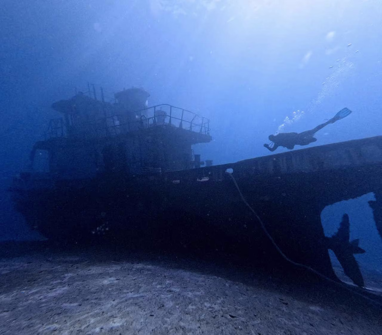 wreck dive 1 (1) Scuba diver exploring a large shipwreck on the seabed, with the dark wreck silhouetted against the deep blue water.